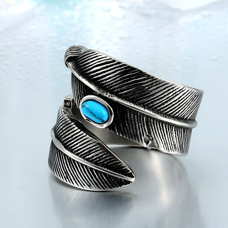 Anel masculino Pluma Turquesa em aço prata com pedra turquesa, exibido em fundo azul claro para destacar o design robusto de penas.