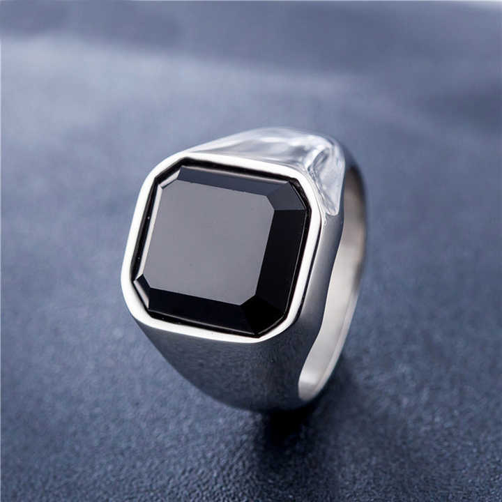 Anel masculino quadrado com pedra preta lapidada em aço, mostrado de um ângulo lateral em fundo azul para realçar a lapidação da pedra e o design sofisticado