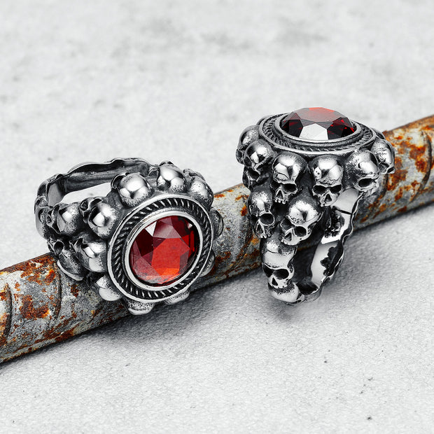 Dois anéis masculinos em aço com crânios e pedra vermelha, posicionados juntos com fundo branco, escorados em um galho de árvore