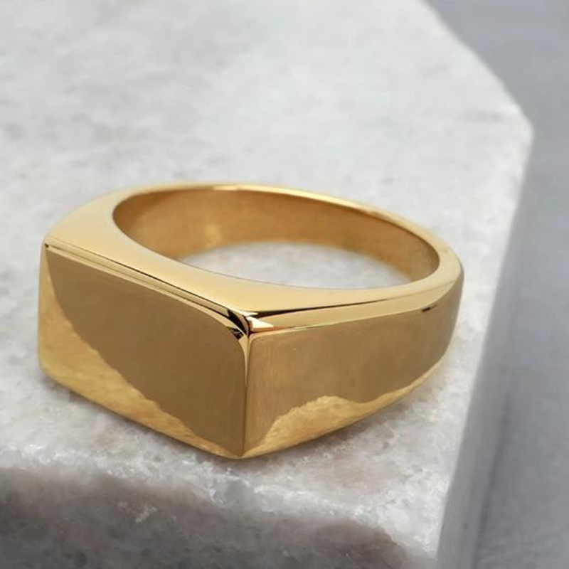Anel masculino Flat Clássico em aço inoxidável dourado posicionado sobre uma pedra branca, criando um contraste que valoriza seus detalhes.