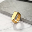 Anel masculino Flat Seta em aço inoxidável dourado com fundo branco e cinza, capturado em um ângulo que evidencia seu formato retangular e o detalhe em seta.