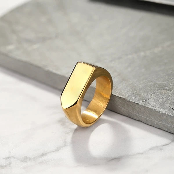 Anel masculino Flat Seta em aço inoxidável dourado com fundo branco e cinza, capturado em um ângulo que evidencia seu formato retangular e o detalhe em seta.