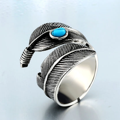 Anel masculino Pluma Turquesa em aço prata, com detalhe de penas e pedra turquesa, mostrado em ângulo lateral sobre fundo azul claro