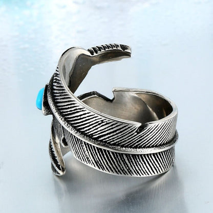 Anel masculino Pluma Turquesa em aço prata, exibido em outro ângulo em fundo azul claro, destacando a pedra turquesa e o design de penas
