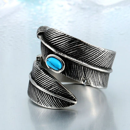 Anel masculino Pluma Turquesa em aço prata com pedra turquesa, exibido em fundo azul claro para destacar o design robusto de penas.