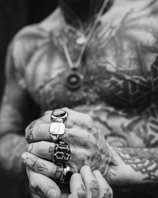 Pessoa com tatuagens usando o anel masculino Número Treze em aço preto, combinado com outras joias, mostrando um visual ousado e estiloso