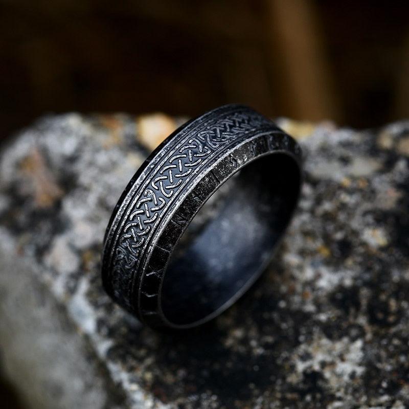 Anel masculino Guerreiro Viking em aço na cor preto antigo, sobre um fundo rochoso cinza, mostrando a textura rústica e o visual envelhecido da peça.