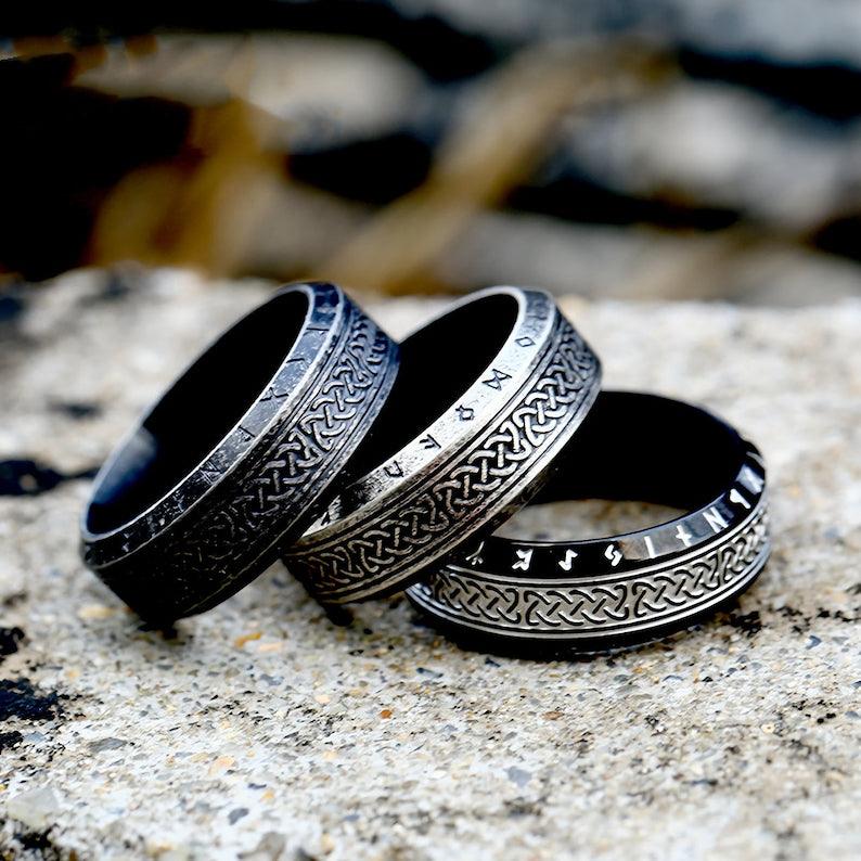 Três anéis masculinos Guerreiro Viking em aço, nas cores preto, preto antigo e prata antiga, juntos sobre um fundo rochoso cinza, realçando os detalhes do design.