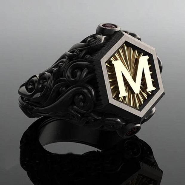 Anel masculino preto maçônico esculpido com a letra "M", capturado em fundo cinza escuro e destacado por seu design detalhado.