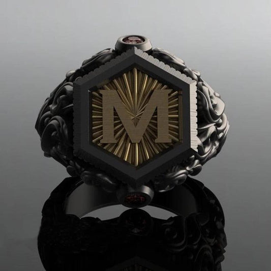 Anel masculino preto maçônico esculpido, exibido em fundo cinza escuro, mostrando o design gravado da letra "M" de um ângulo diferente.