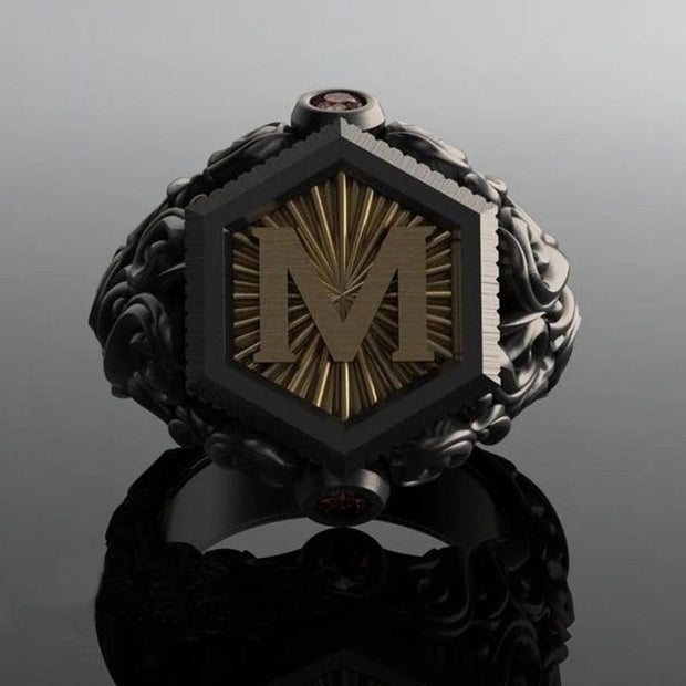 Anel masculino preto maçônico esculpido, exibido em fundo cinza escuro, mostrando o design gravado da letra "M" de um ângulo diferente.