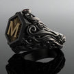Anel masculino preto maçônico esculpido, visto de um quarto ângulo com fundo cinza escuro, destacando o acabamento e a estrutura elegante.