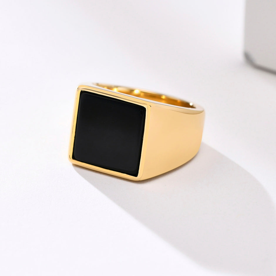 Anel masculino quadrado em aço dourado com pedra preta lisa, exibido em fundo claro para destacar o contraste entre o dourado e a pedra preta