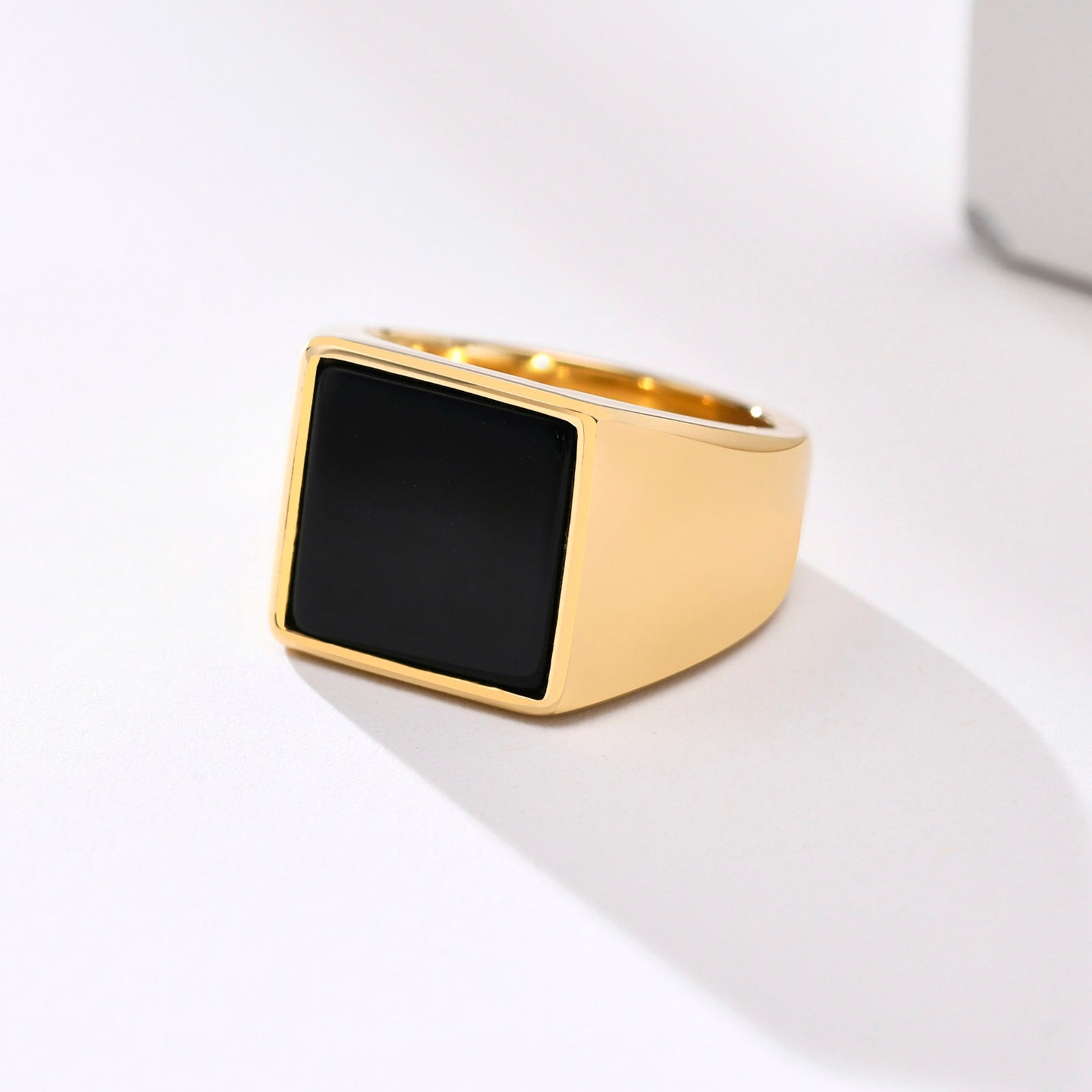 Anel masculino quadrado em aço dourado com pedra preta lisa, exibido em fundo claro para destacar o contraste entre o dourado e a pedra preta