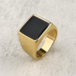 Anel masculino quadrado em aço dourado com pedra preta lisa, mostrado de um ângulo lateral em fundo claro para destacar o brilho do metal.