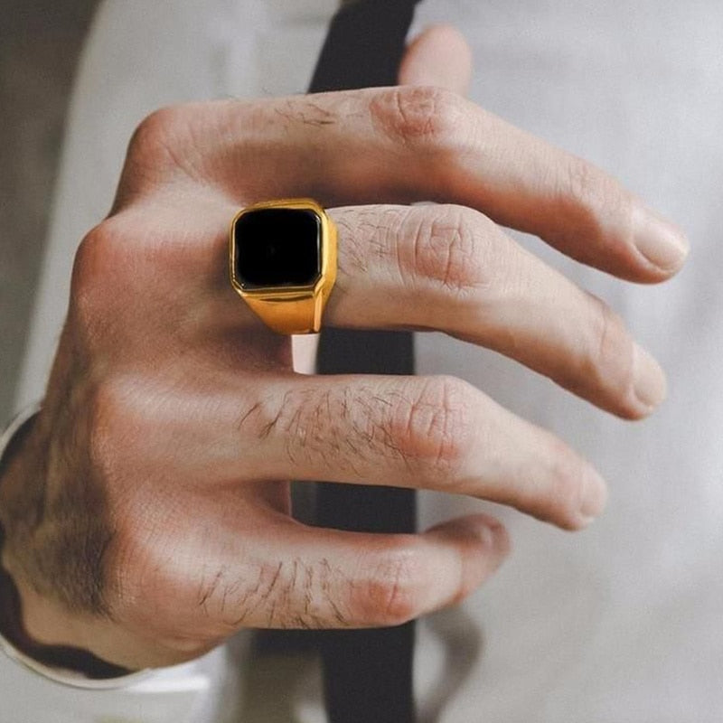 Anel masculino quadrado em aço dourado com pedra preta lisa, sendo usado no dedo, exibindo o contraste de cores e a elegância do anel.