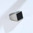 Anel masculino quadrado em aço prata com pedra preta lisa, exibido em fundo claro para destacar o contraste elegante entre a pedra e o metal.