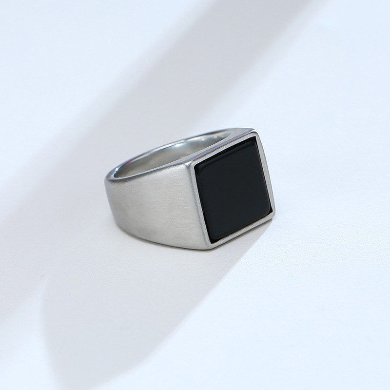 Anel masculino quadrado em aço prata com pedra preta lisa, exibido em fundo claro para destacar o contraste elegante entre a pedra e o metal.