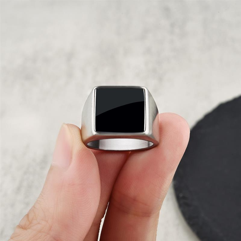 Anel masculino quadrado em aço prata com pedra preta lisa, sendo segurado na ponta dos dedos para exibir os detalhes refinados do design.