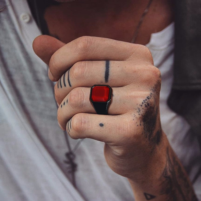 Anel masculino quadrado com pedra vermelha em aço preto, sendo usado no dedo para mostrar seu estilo robusto e o ajuste