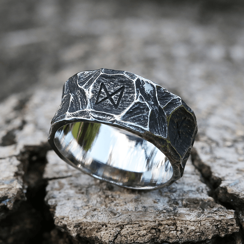 Anel masculino Viking rústico em aço prata, mostrado de outro ângulo sobre fundo rochoso claro, evidenciando o acabamento rústico e o design diferenciado.