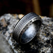 Anel masculino Guerreiro Viking em aço na cor prata antiga, exibido sobre um fundo rochoso cinza, destacando os detalhes e a cor envelhecida do anel.